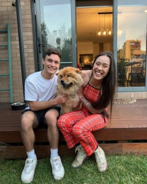 A young woman and man sit on the step of what looks like a wooden patio outside a warm and inviting home; both are smiling and holding a furry chow chow dog between them. 