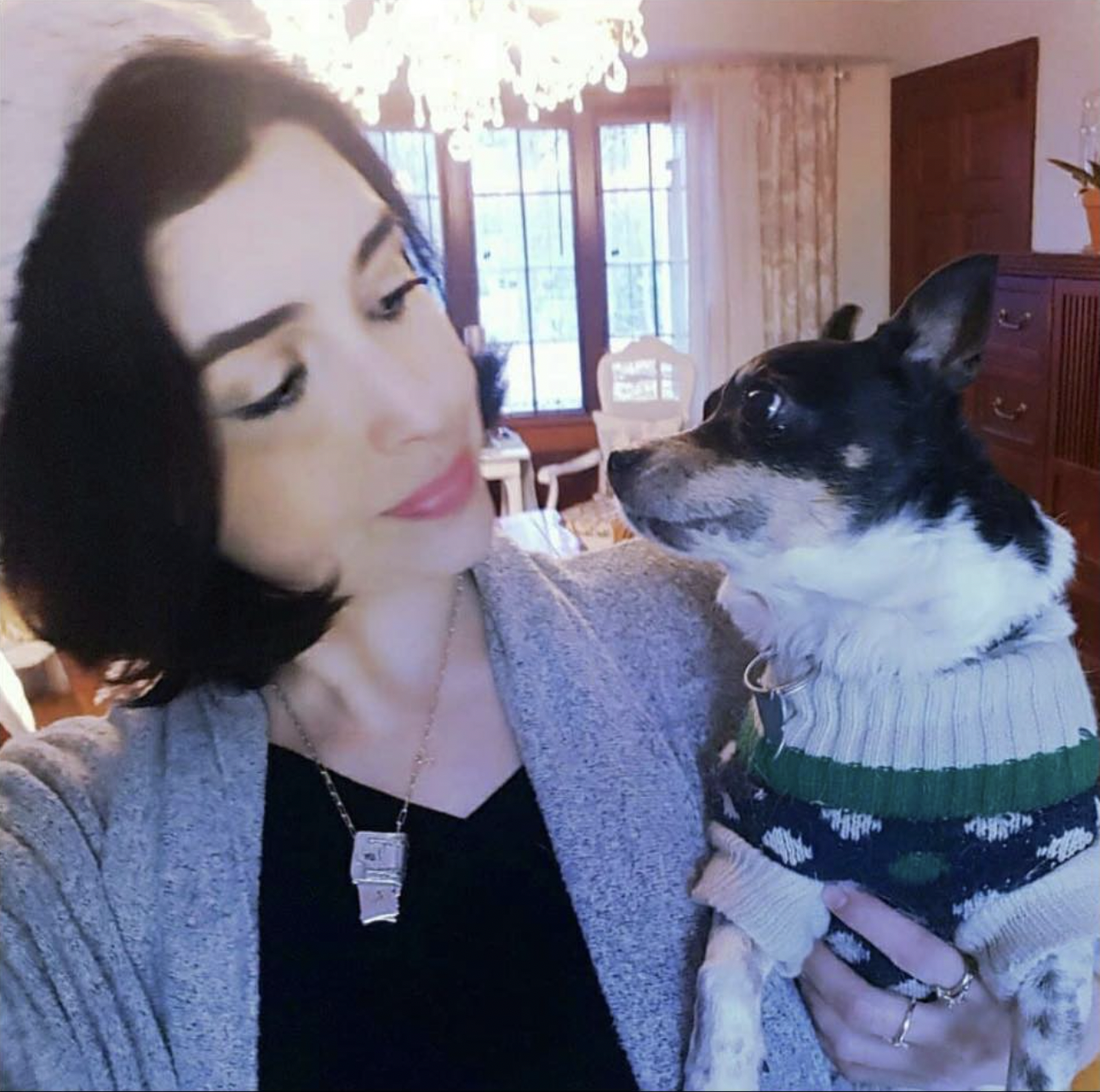 A woman with black hair looks at the dog she's carrying at right. She wears a gray sweater over a black shirt with a necklace; the dog, with a largely black-haired face and what seems to be a white-haired muzzle and body, is in a sweater of green, blue, and white.