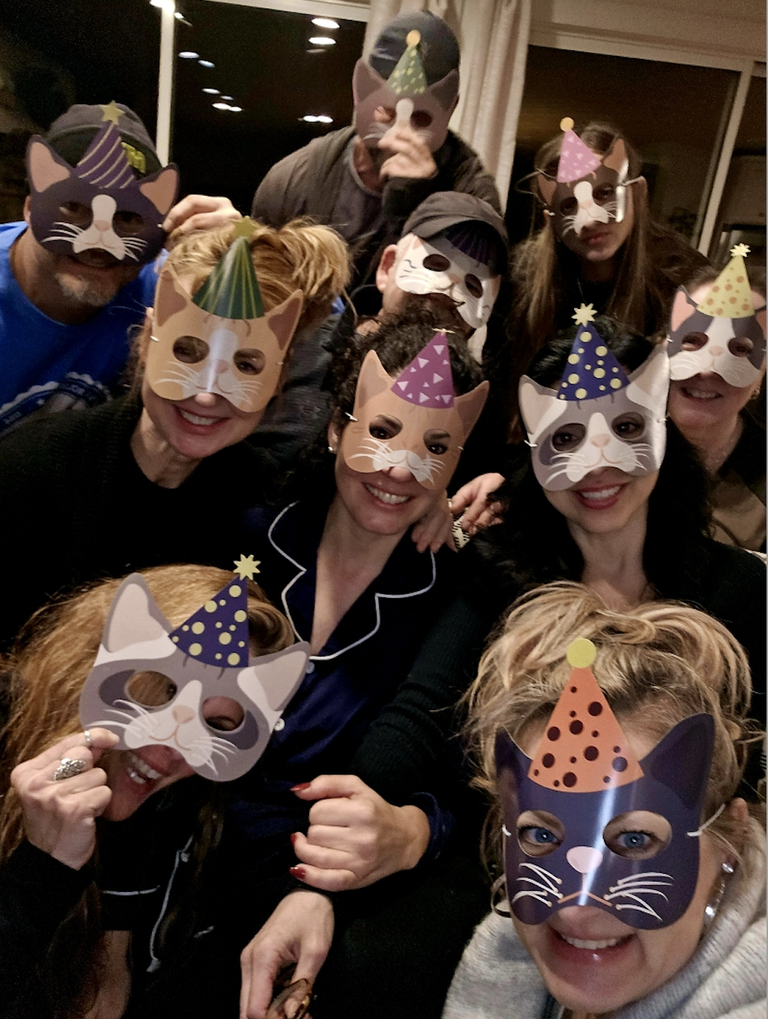 A group of 10 people cram into the frame of a selfie. They're all wearing cat-themed party masks, so we can't see their faces. Some are women and some are men. 