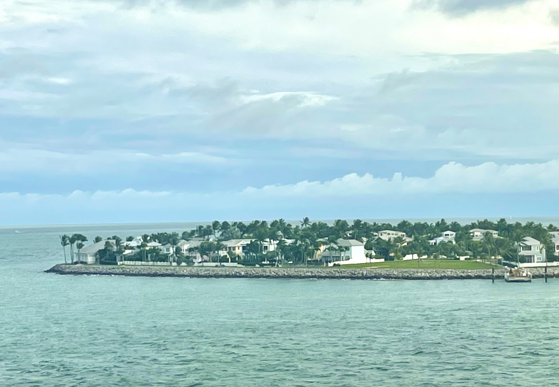 A photo taken from a ship on the ocean shows a tropical island filled with palm trees and houses. The day is partly cloudy and appears windy.