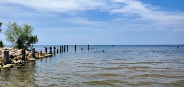 A photo taken from the beach shows a large lake extending toward a clear horizon. On the left, rocks extend out into the water, along with a number of wooden posts.