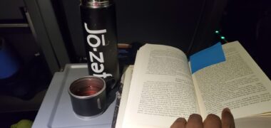 A photo shows a thermos and a book resting on a folding lap desk, with a person's hand holding the book open.