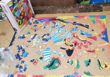 Very colorful pieces of a jigsaw puzzle are laid out on a piece of cardboard. The puzzle box is standing upright in a corner of the cardboard. 